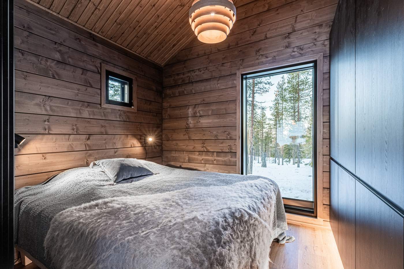 Bedroom with timber walls, pendant light and a large window onto the forest